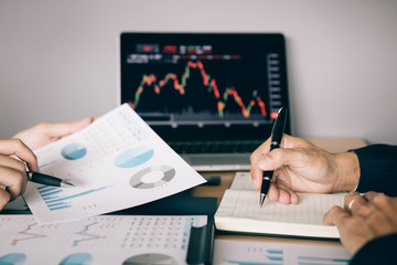 Two investors are working together with analyzing the stock data graphs in the paper and viewing the data on the laptop screen.