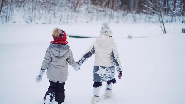 Back view of two little children walking in the snow while holding hands together. Children in warm clothing have a walk near the forest on the background of white snow. Slow motion.