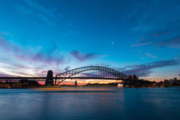 Boat trail passing through Sydney Harbour Bridge at dusk.