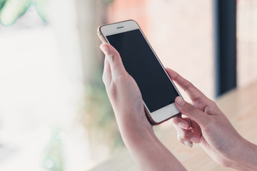 Croppes shot women hand holding white smartphone with blank screen on wooden counter bar in cafe. vintage tone for business technology background concept. closeup mobile.