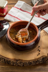 Female hand holding spoon with forest mushroom soup with potatoes in clay pot on wooden table