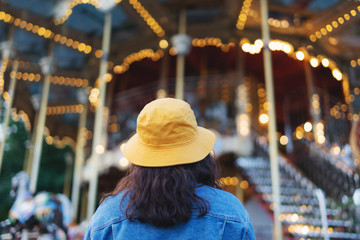 Young woman in yellow hat looking at roundabout in theme-park in summer