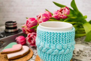 A white cup of tea in a knitted blue case with an old camera and pink flowers tulips and sweets, gingerbread on a textural table. Spring, winter concept