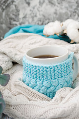 White mug with tea in a knitted blue frame and wrapped in a white knitted plaid. In the background, sprigs of eucalyptus and cotton. Breakfast in bed. Cozy. Spring, winter.