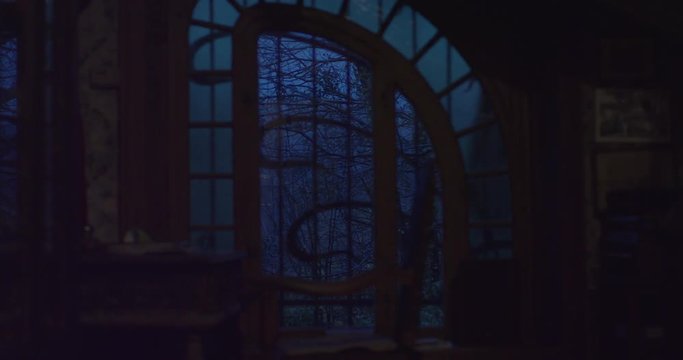 Old creepy window of a scary haunted house overlooking a haunted forest with trees moving in the wind. Old objects in old solid wood, old wallpaper damaged, changing focus. Shot on red epic 6K.