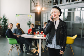 Caucasian pretty business woman in hat and jacket using phone. successful European business woman, talking on the phone, standing on background of colleagues on meeting in modern loft office