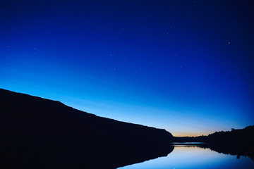 blue night sky after sunset on the background of the silhouette of a hill photographed from the shore of a calm mirror lake