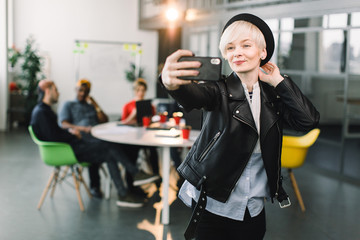 Smiling blond business woman in black leather jacket and hat making selfie on phone at small office, young workers freelancers have a meeting and discussion at the table