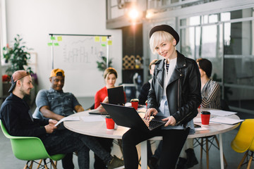 Multiethnic diverse group of happy colleagues working together in modern loft office. Creative young girl designer in hat and leather jacket working on laptop and sitting on the table
