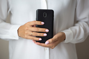 Close-up photo of elegant light pink manicure over white shirt background, tender women's hands with perfect nails hold a mobile phone