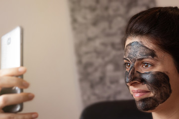 brunette italian girl with a black beauty mask on her face taking a selfie. natural light coming from the window in front of her.