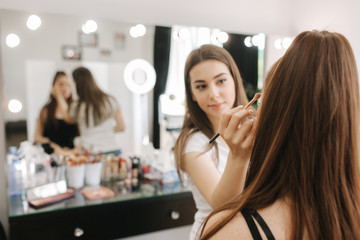 Beautiful femalemakeup artist doing makeup for a young redhead girl in a beauty salon sitting in front of a large mirror. Concept of preparation for the holiday and meeting