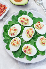 Deviled eggs stuffed with avocado, egg yolk and mayonnaise filling, garnished with bacon, on spinach leaves, vertical top view