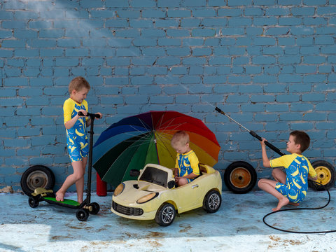 Funny children in boys' bathing suits posing in front of camera with high-pressure car wash and toy yellow car and scooters. Brothers are trying to wash car. Rainbow umbrella
