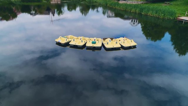 Drone view of lake, which reflects cloudy sky, and yellow paddleboats floating on it. 4K.