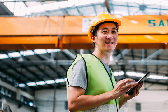 Young Asian male heavy industral worker holding a digital tablet inside manufacturing and factory site with crane behind