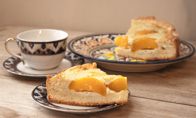 Slice of peach apricot pie or cheese sour cream cake on bright saucer with tea cup on wooden table