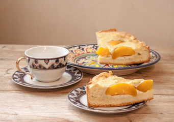 Slice of peach apricot pie or cheese sour cream cake on bright saucer with tea cup on wooden table