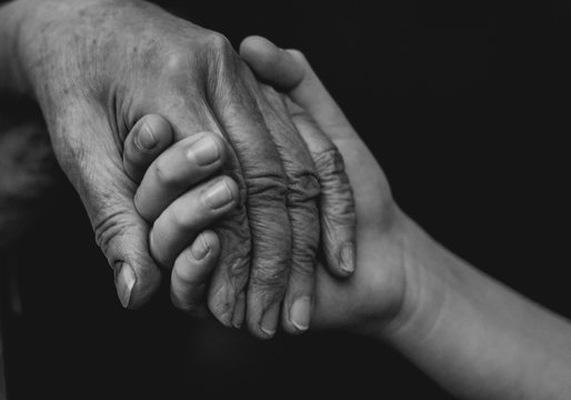 The hands of an old man and a child hold each other against a black background.