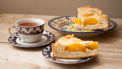 Slice of peach apricot pie or cheese sour cream cake on bright saucer with tea cup on wooden table