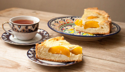 Slice of peach apricot pie or cheese sour cream cake on bright saucer with tea cup on wooden table