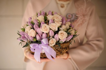 Girl in the coat holding a wicker basket of tender creamy color roses and violet tulips
