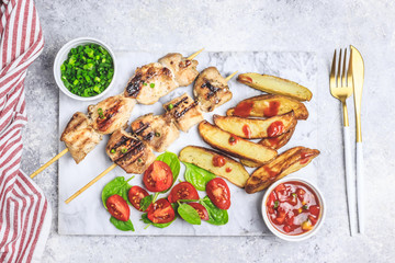 Grilled chicken skewers with baked potato, green onion and salsa sauce on a marble plate on a light gray background. Top view.