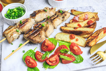 Grilled chicken skewers with baked potato, green onion and salsa sauce on a marble plate on a light gray background. Selective focus