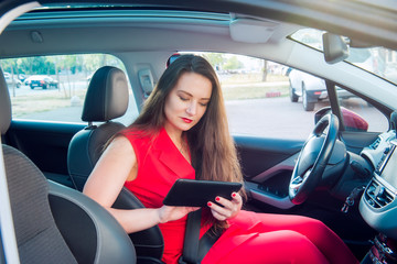 Portrait of serious business lady, caucasian young woman driver in red summer suit setting up the route on a navigator while sitting behind the wheel car. Side view. Selective focus, copy space.