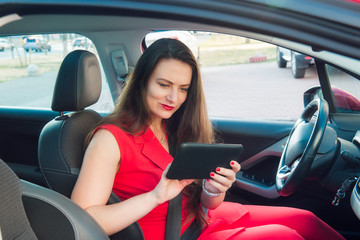 Portrait of confused and anxious business lady, caucasian young woman driver in red summer suit setting up the route on a navigator while sitting behind the wheel car. Selective focus, copy space.