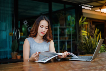 Close up shot of a young female university student writes a note and assignment with a laptop beside outside the cafe with blur green tree background. Students self learning outside the campus.