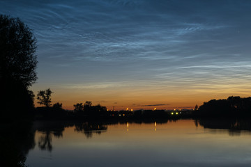 Noctilucent clouds over northwestern Europe, seen from the shore of a lake at night on a calm night