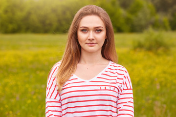 Outdoor portrait of happy smiling girl with long straight hair in white shirt with red stripes posing in summer meadow, has calm and pleasant facial expressions, being photographed by friend.