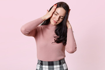 Obraz na płótnie Canvas Studio shot of attractive young woman with terrible headache, keeps both hands on head, needs to take medicine, brunette female in rose shirt and checkered skirt, isolated over pink background.
