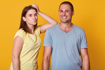 Fototapeta premium Photo of smiling cheerful man having pleasant facial expression, looking directly at camera. Girlfriend putting her arm on man, looking funny, wearing casual t shirt, isolated over yellow background.