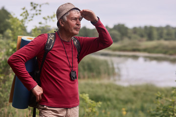 Obraz premium Photo of caucasian man hipster man with backpack and rug in nature background. Relax time on holiday, eldery male looking far away, dressed casual red shirt and cap. Traveling and active rest concept.