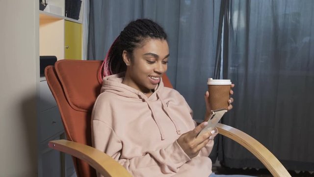 Young black girl sitting in Chair in her room and holding smartphone in hands and chatting, while drinking coffee. This video depicts social networks, instagram, Facebook, browsing web, searching.