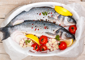 Two raw fresh sea bass ready for baking with tomatoes, lemon, red onion, garlic, ginger, parsley and spices. Prepare a tasty and healthy lunch or dinner in a Mediterranean style. Selective focus