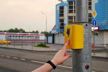 The hand of a girl with a clock presses the button of the pedestrian crossing. The button is lit in...
