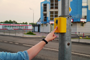 The hand of a girl with a clock presses the button of the pedestrian crossing. The button is lit in red, electronic pedestrian crossing. A hand sign indicates a button.
