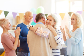 Happy excited mature lady with gray hair holding birthday gift and hugging friend while thanking him for gift, other friends clapping hands