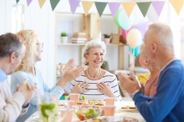 Group of best friends sitting at dining table and applauding happy retired lady while...