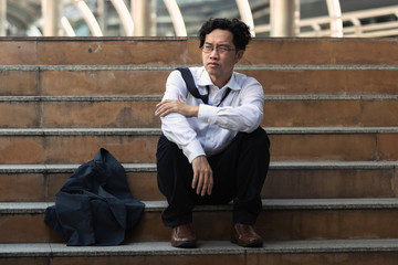 Obraz premium Depressed stressed young Asian business man in suit with hands on head sitting on stairs. Unemployment and layoff concept.