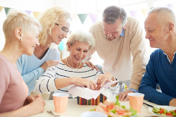 Jolly positive senior woman with curly hair sitting at table and opening gift box in excitement, her friends supporting her