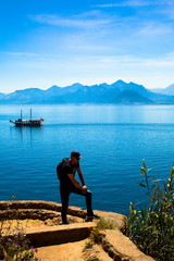 Naklejka premium Young man stands on a rock on the background of a beautiful view of the Mediterranean Sea, the mountains and the ship. Turkey, Antalya.