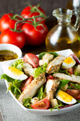 Fresh Caesar salad made of tomato, ruccola, chicken breast, eggs, arugula, crackers and spices. Organic ingredients in a white, transparent bowl on wooden background