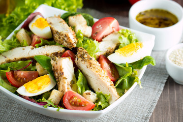 Fresh Caesar salad made of tomato, ruccola, chicken breast, eggs, arugula, crackers and spices. Organic ingredients in a white, transparent bowl on wooden background