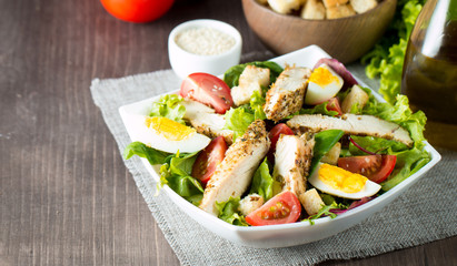 Fresh Caesar salad made of tomato, ruccola, chicken breast, eggs, arugula, crackers and spices. Organic ingredients in a white, transparent bowl on wooden background