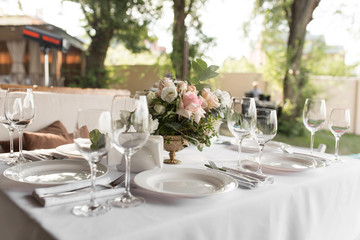 Wedding table setting decorated with fresh flowers in a brass vase. Wedding floristry. Banquet table for guests outdoors with a view of green nature. Bouquet with roses, eustoma and eucalyptus leaves