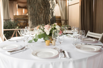 Wedding table setting decorated with fresh flowers in a brass vase. Wedding floristry. Banquet table for guests outdoors with a view of green nature. Bouquet with roses, eustoma and eucalyptus leaves